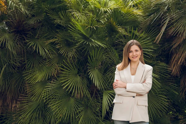 积极的女企业家 棕色长发 穿着时髦的米黄色西服 微笑着 看着镜头 站在绿油油的热带植物旁边 两臂交叉 — 图库照片