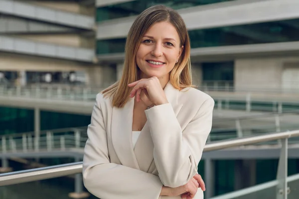 Positive young female executive manager in smart casual suit touching chin while standing near railing outside modern office building looking at camera