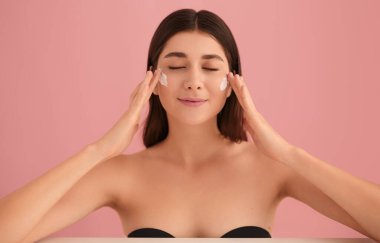 Siyah straplez sutyenli narin genç esmer yanağına beyaz krem sürerken pembe arka planda gözleri kapalı yüz derisine özen gösteriyor.