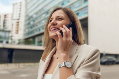 Resmi kıyafetleri içinde uzun sarı saçlı neşeli yetişkin iş kadını mutlu bir şekilde gülümsüyor ve uzaklara bakıyor, cep telefonuyla konuşurken modern binaların yakınında duruyor.
