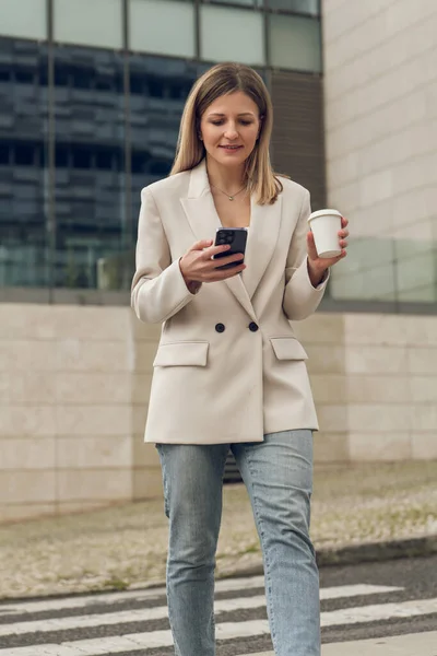 Happy young female manager with long blond hair in smart casual outfit, smiling and messaging on smartphone while crossing road with cup of takeaway coffee during break