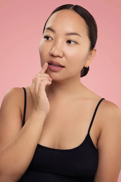 Attractive ethnic young female in black top touching lip while standing against pink background looking away