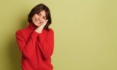 Kırmızı balıkçı yakalı, elleri kenetlenmiş, gülümseyen ve canlı arka planda kameraya bakarken yanağına dokunan pozitif, genç bir kadın.