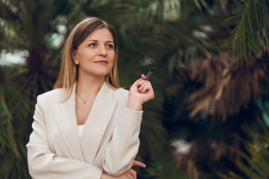 Yeşil parktaki ağaç yapraklarının üzerindeki fotokopi alanını gösterirken zarif kıyafetli, gülümseyen ve bakışlarını çeviren hoş bir kadın.