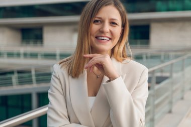 Gülümseyen, sarı saçlı, bej ceketli, iş merkezinin balkonunda duran ve çenesine dokunurken kameraya bakan yetişkin bir girişimci.