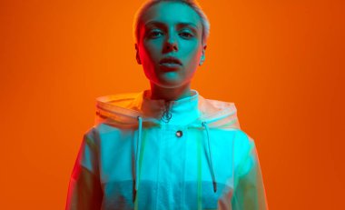 Ekin, genç bayan milenyum gömleği, şeffaf yağmurluklu sarı saçlar, neon stüdyodaki turuncu arka planda kameraya bakıyor.