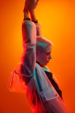 Kısa sarı saçlı, şeffaf yağmurluklu, turuncu arka planda duran, kollarını kaldırmış ve neon ışıklarla gözlerini kaçıran kendine güvenen genç kadın modelin yan görüntüsü.