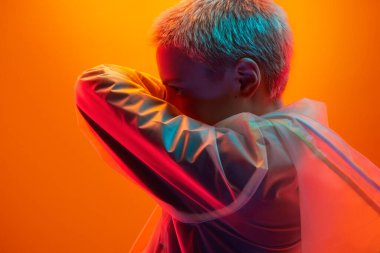 Ciddi, sarışın, şeffaf yağmurluklu, yüzü kolla kaplı ve neon stüdyodaki turuncu arka plana bakan genç bir kadının yan görüntüsü.