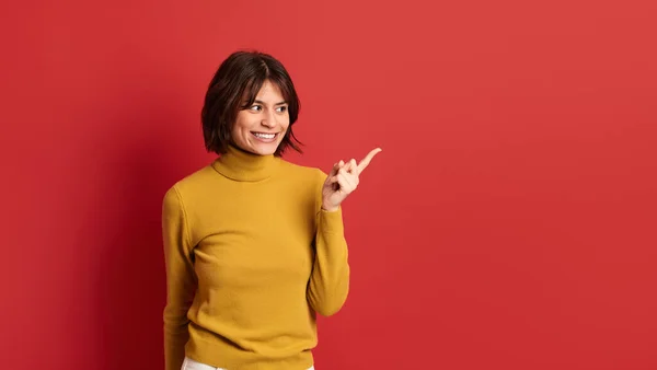 幸せな若いです女性で黄色のセーター笑顔と横に見て 指を指している間にコピースペースに対して赤 — ストック写真
