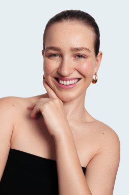 Siyah saçlı, parlak tenli, neşeli genç bir manken yüzüne dokunuyor ve beyaz arka planda kendine güvenen kameraya bakıyor.