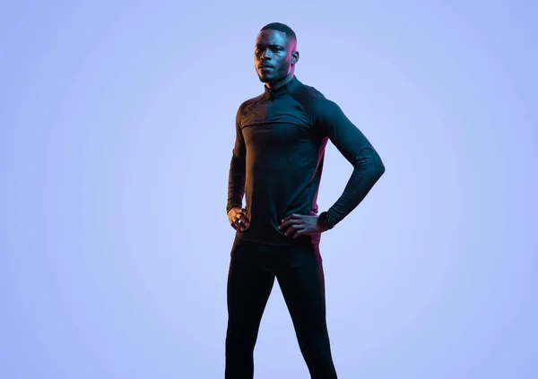 身穿黑色运动衫和腿的认真的非裔美国男性运动员 手挽腰站在蓝色的背景上 看着镜头 — 图库照片