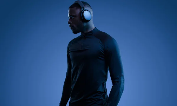 身穿黑色运动服的有自信的年轻非洲裔美国运动员 穿着现代无线耳机听音乐 背向远方看蓝色背景 — 图库照片