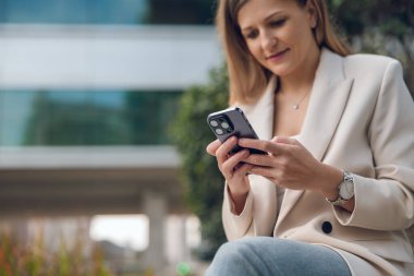 Kendine güvenen, uzun saçlı, şık spor ceketli bir girişimci akıllı telefonuyla mesajlaşırken modern ofis binasının yanındaki bankta oturuyor.