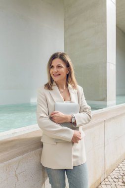 Havuz kenarında gün ışığında taştan kenarları olan yüzme havuzuna bakarken elinde netbook tutan şık kıyafetli neşeli bir kadın.