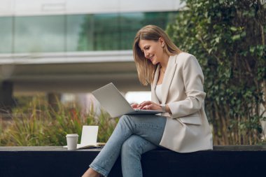 Spor ceketli, spor ceketli bir kadının park bankında kahve alıp uzaktan çalışırken dizüstü bilgisayarda yazışının yan görüntüsü.