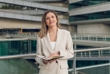 Tarz sahibi bej takım elbiseli, uzun sarı saçlı, pozitif iş kadını gülümseyen ve kameraya bakan, modern ofis binasının önündeki not defterinde duran,