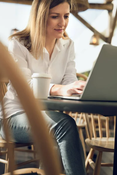 紙コップでコーヒーを飲みながらノートパソコンでタイプしながら カフェから離れた場所で働いている間 カジュアルな服で大人の女性のフリーランサーを笑顔 — ストック写真