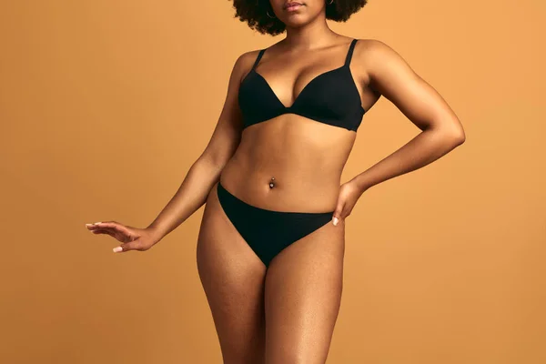 Elegant African female in black underwear with belly piercing and arm outstretched standing against orange background