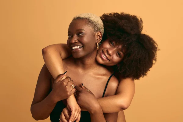 Happy African female with Afro hair embracing girlfriend from behind while standing against orange background having fun