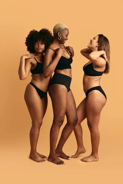 Full body of multiracial women in black lingerie standing in studio touching shoulders fixing hair and adjusting bra against orange background