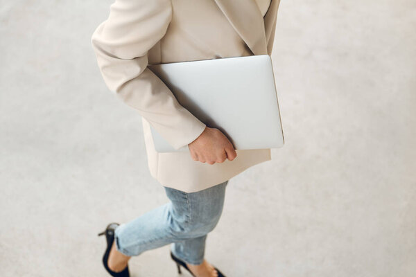 Сверху неузнаваемая стильная женщина-менеджер в бежевых джинсах и туфлях на высоком каблуке, ходит по улице с ноутбуком в руке в дневное время