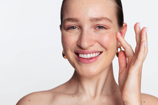 Closeup portrait of positive young female with perfect skin and natural makeup smiling and looking at camera while touching face on white background