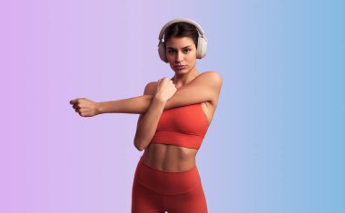 Kendine güvenen genç İspanyol bayan sporcu modaya uygun spor kıyafetleriyle kablosuz kulaklıkla müzik dinliyor ve arka planda trisepsleri esnetirken kameraya bakıyor.