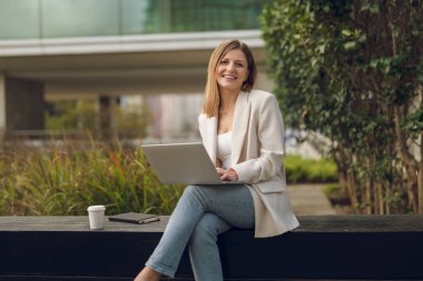 Zeki gündelik kıyafetli genç bayan iş kadını yeşil çalıların yanındaki bankta oturmuş elinde bir fincan sıcak içecek ve planlayıcıyla Netbook kullanarak uzaktan çalışıyor.