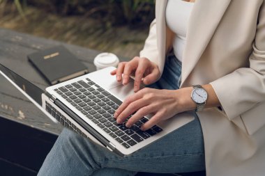 Yüksek ekin açılı, sıradan şık giyinen, dizüstü bilgisayarla bankta oturan ve uzak bir proje üzerinde çalışan anonim bir kadın.