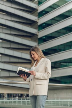 Modern ofis binasının yanında dikilirken, uzun sarı saçlı, şık kıyafetli, defter okuyan, konsantre genç bayan girişimcinin yan görüntüsü.