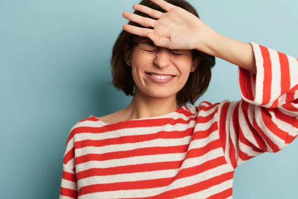 Portrait of smiling woman with short hair in striped sweater standing near blue wall and hitting forehead realizing failure
