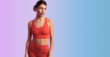 Kırmızı aktif giysili İspanyol kadın modeli dikkat çekmeden arkaplanda dikilirken diğer tarafa bakıyor.