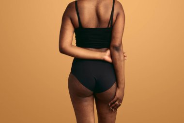 Siyah iç çamaşırlı Afro-Amerikan kadın modelin bej arka planda durduğunu ve elleri arkada durduğunu gördüm.