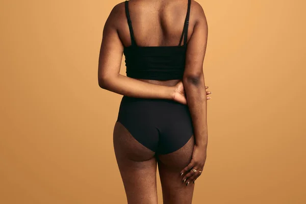 Back view of unrecognizable African American plus sized female model in black underwear standing against beige background with hands behind back