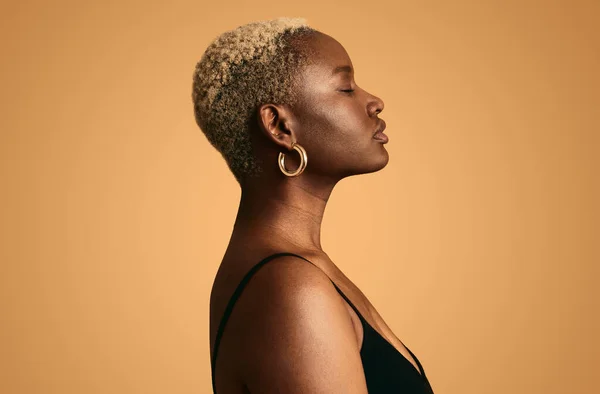 Side view of pensive young African American female model in black top with short hair and earrings standing against beige background with closed eyes