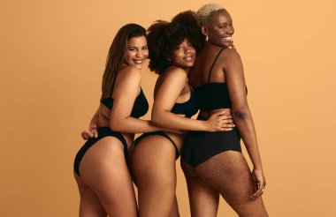 Siyah iç çamaşırı giyen, birbirine yakın duran, turuncu arka plan kavramına dokunan, gülümseyen, çok ırklı genç kız arkadaşlar.