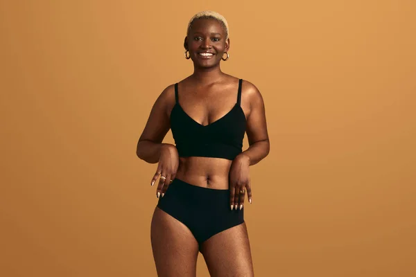 Positive self assured black female with short blond Afro hair touching panties while standing against orange background concept of body confidence and self acceptance