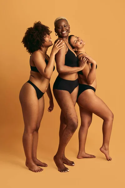 Full body of happy young multiracial female models in black underwear standing against yellow background and smiling while touching each other in studio