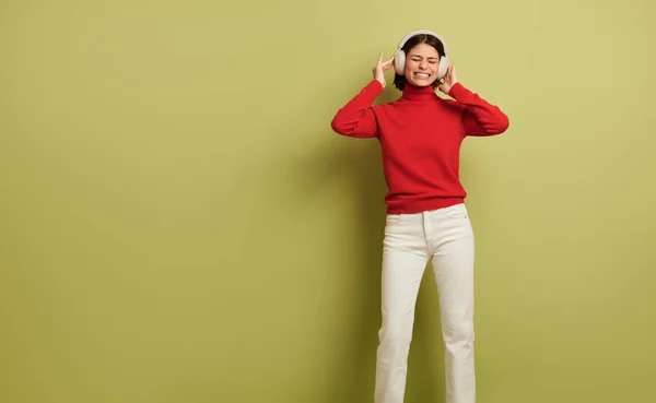 明るい若いですヒスパニック系の女性の千年紀で暗い髪で赤いセーターと白いジーンズ 緑の背景にワイヤレスヘッドフォンで音楽を聴きながら 目を閉じて幸せに笑顔 — ストック写真