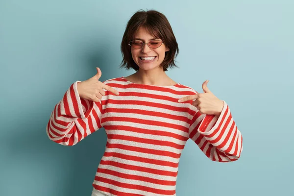 감넘치는 긍정적 모델붉은 줄무늬 스웨터와 선글라스를 웃으며 배경에 자신을 가리키며 — 스톡 사진