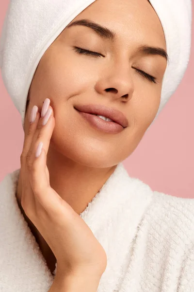 身着白色浴衣 头戴毛巾 面带微笑的年轻女性 紧闭双眼 在粉红背景的美容活动中使用护肤产品 — 图库照片