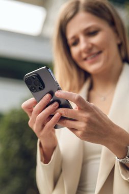 Çevrimiçi sohbet ederken akıllı telefon kullanan, şık giyinmiş, düşük açılı gülümseyen genç bir kadın ekranı izliyor.