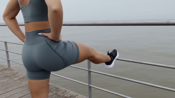 流行のスポーツウェアと良い物理的な形のストレッチとウォーミングアップの認識できない運動女性のバックビュー海沿いの遊歩道で屋外でトレーニングする前に — ストック動画