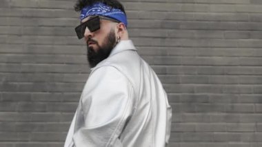 Güneş gözlüklü ve saç bantlı genç sakallı adam hip hop ve sokak kültürü tarzında arkasını dönüp kameraya bakıyor.