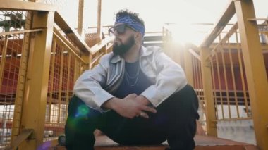 Sokak kıyafetleri ve güneş gözlüğü takan şık sakallı adam kameraya sokak jesti yapıyor. Basamaklarda havalı bir break dansçının kasıla kasıla oturması..