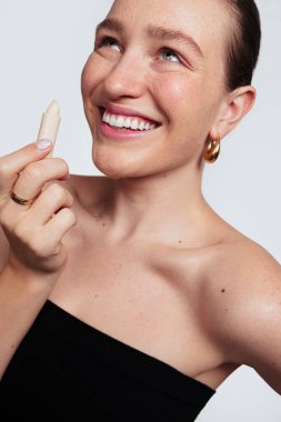 Kendine güvenen, çilli, kahverengi saçlı, gülümseyen ve beyaz arka plana dudak kremi sürerken kafasını kaldıran genç bir model.