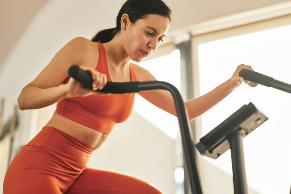 Low angle of fit young athletic female in sportswear training muscles on elliptical trainer during fitness workout at gym