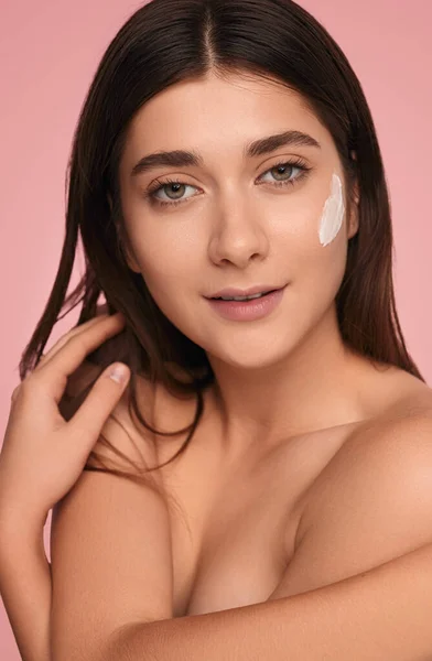 Self assured young topless female model with long dark hair and facial cream on cheek, covering breast with arms and looking at camera against pink background