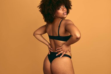 Afro saçlı, siyah iç çamaşırı giyen, elleri belinde, turuncu arka planda kameraya bakan Afro-Amerikan bir kadın.