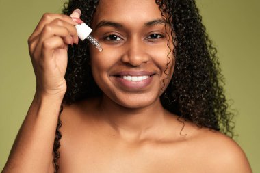 Kıvırcık saçlı genç bir kadın yeşil arkaplandaki kameraya bakarken yüzüne serum sürüyor.